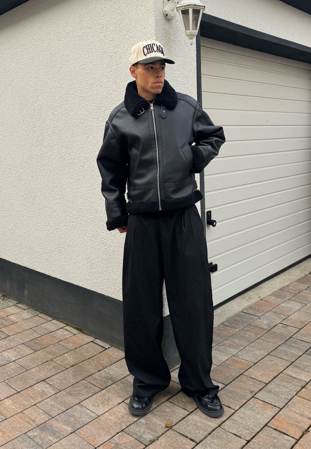 Blouson aviateur en cuir noir avec col en peau retournée, associé à un pantalon large noir à fines rayures et des chaussures noires, accompagné d'une casquette de baseball crème.