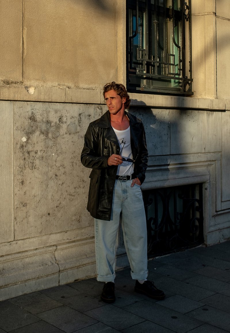 Juodos odinės ilgos striukės, uždengiančios baltą marškinėlį, šviesiai mėlynos plačios džinsinės kelnės ir juodi batai. Laiko saulės akinius, stovėdamas prie tekstūruoto sienos.