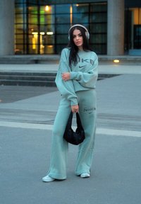Mintgrønn Nike sweatshirt og matchende joggebukser med store logo-detajer. Modellen holder en svart bag og har på seg sølvhodetelefoner. Casual gatekultur.