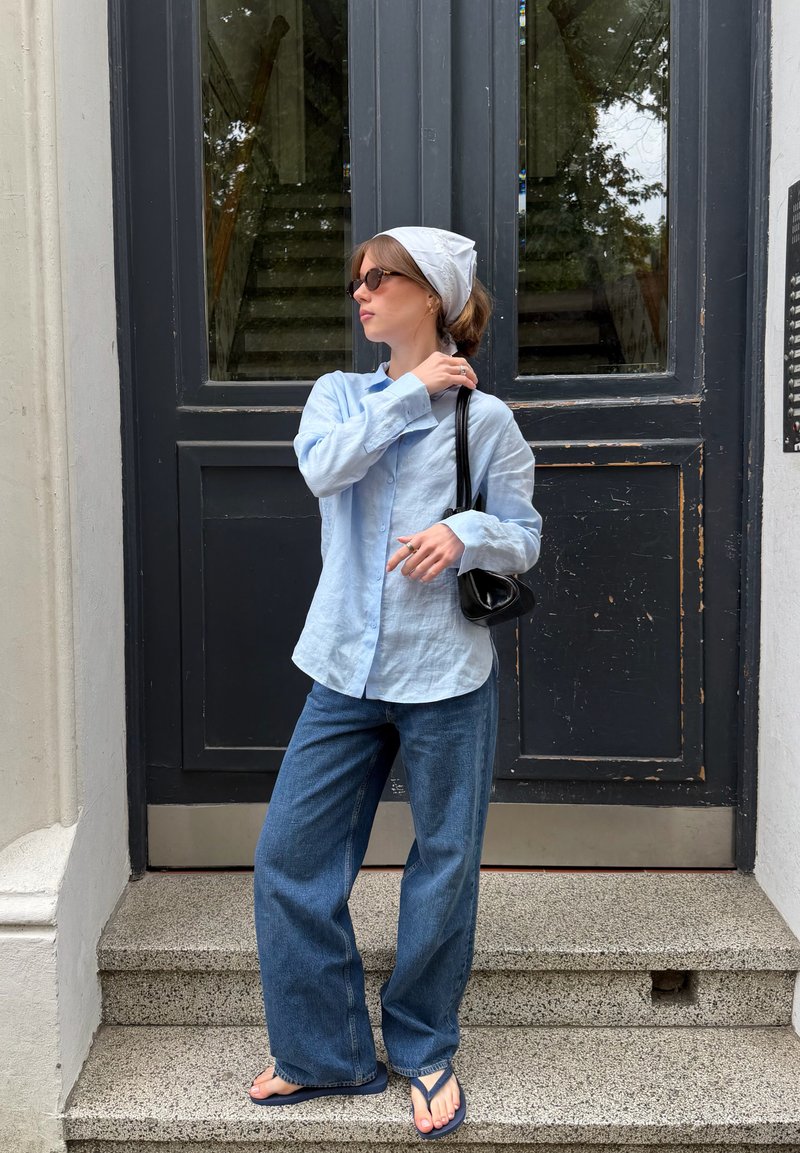 Femme portant une chemise bleu clair, un jean large, un foulard blanc sur la tête, des lunettes de soleil et des tongs, debout sur des marches en pierre devant des portes doubles noires.