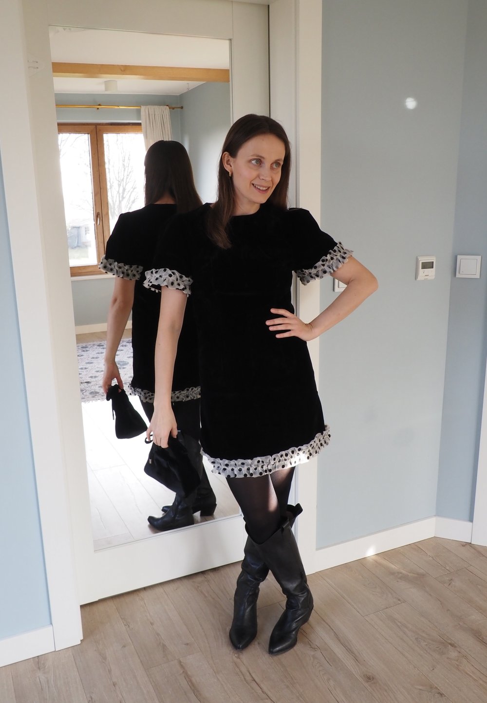 Robe en velours noir avec manches bouffantes, ourlet orné de pois, associée à des bottes montantes noires et un petit sac à main, reflétée dans un miroir.