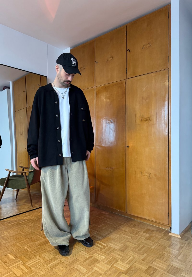 Άνδρας που φοράει μαύρο καπέλο, μαύρο πουκάμισο με κουμπιά, λευκή μπλούζα και φαρδιά μπεζ παντελόνια, στέκεται σε δωμάτιο με ξύλινο πάτωμα, καρέκλα και μεγάλο ξύλινο ντουλάπι.