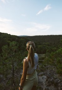 O persoană stă pe un stâncă cu vedere spre o vale verde, purtând un top ușor și pantaloni scurți deschiși, cu părul lung strâns la spate.
