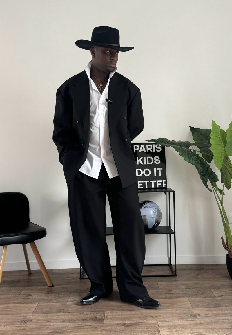 Veste noire à double boutonnage, chemise blanche en dessous, pantalon noir à jambes larges et grand chapeau noir. Chaussures à surface lisse. Cadre intérieur avec des plantes.