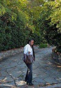 Borsa a mano in pelle marrone con doppi manici, abbinata a una camicia azzurro chiaro e jeans scuri svasati, su un sentiero di pietra circondato da verde.