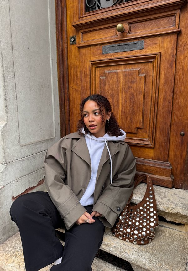 Ung kvinne med krøllete hår sitter på steintrapp ved en tredør, iført en grå jakke, hvit hettegenser, svarte bukser, ved siden av en naglet brun veske.