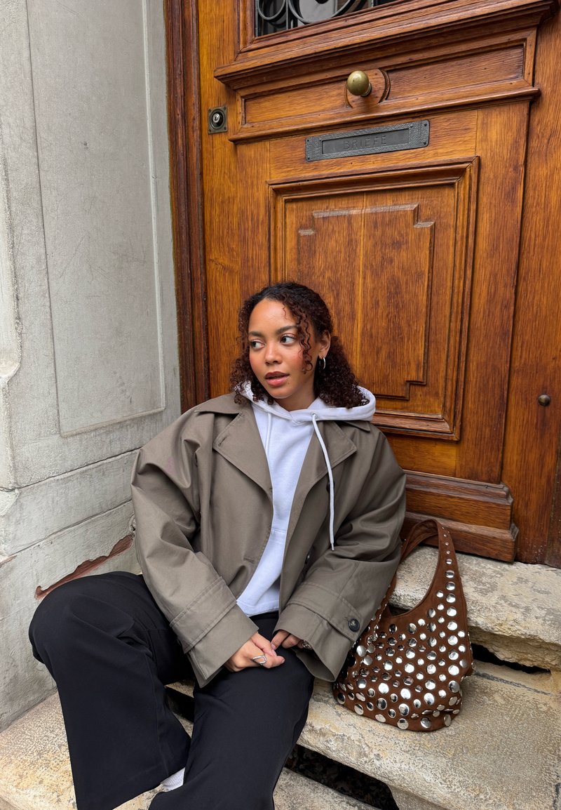 Jeune femme aux cheveux bouclés assise sur des marches en pierre près d'une porte en bois, portant une veste grise, un sweat à capuche blanc, un pantalon noir, à côté d'un sac marron clouté.