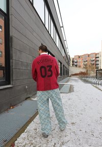 Punane kootud kampsun, millel on Adidas'i bränding ja number 03 seljal, sobitatud helesinine ruuduline pükstega. Väljas, lumikattega maastik.