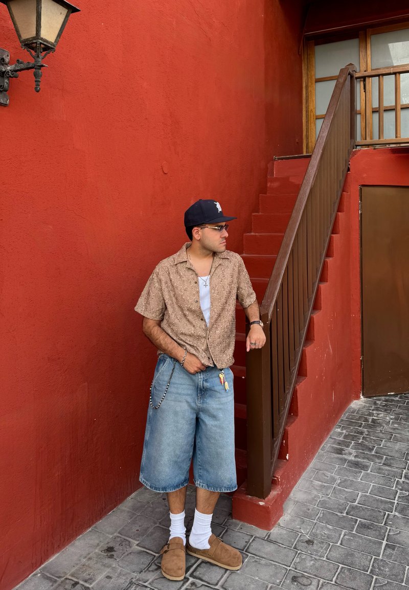 Chemise à manches courtes à motifs bruns, débardeur blanc, shorts en denim bleu clair, sabots beiges, chaîne en argent, contre un mur rouge avec des escaliers.