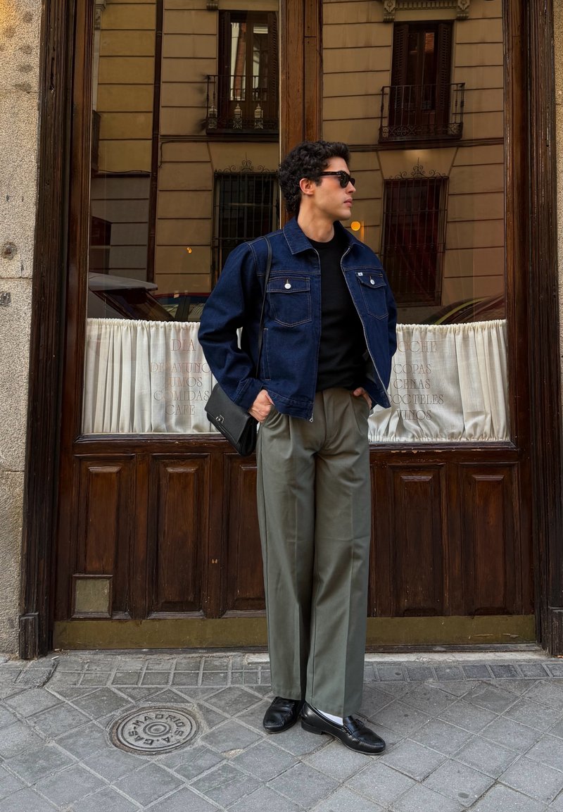 Jeune homme portant une veste en jean foncée, une chemise noire, un pantalon gris, des chaussures noires et des lunettes de soleil, debout les mains dans les poches près d'une porte en bois avec des fenêtres.