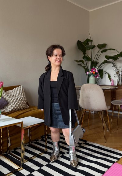 Mujer con blazer negro, falda de mezclilla y botas plateadas de pie en la sala, que tiene una alfombra a rayas, un sofá mostaza y plantas sobre suelo de madera.