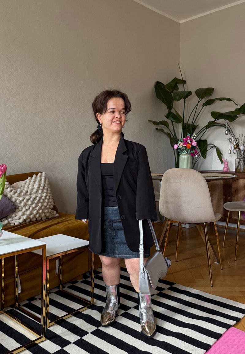 Femme en veste noire, jupe en jean et bottes argentées se tient dans un salon avec un tapis rayé, un canapé moutarde et des plantes sur un sol en bois.