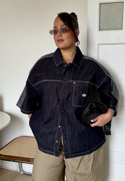 Mujer con chaqueta vaquera oscura de gran tamaño con rayas blancas en las mangas, pantalones beige, gafas de sol, sosteniendo un bolso de mano negro tejido.