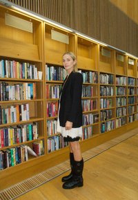 Juodas bleizeris, baltos suknelės su nėriniu, ir juodi iki kelių auliniai batai. Medinės knygų lentynos, užpildytos spalvingomis knygomis. Šilta apšvietimas ir mediniai grindys.