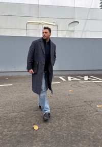 Manteau gris tissé à silhouette longue, pull noir, jean bleu et chaussures foncées. Tenant un dossier noir ; marchant sur une surface pavée.