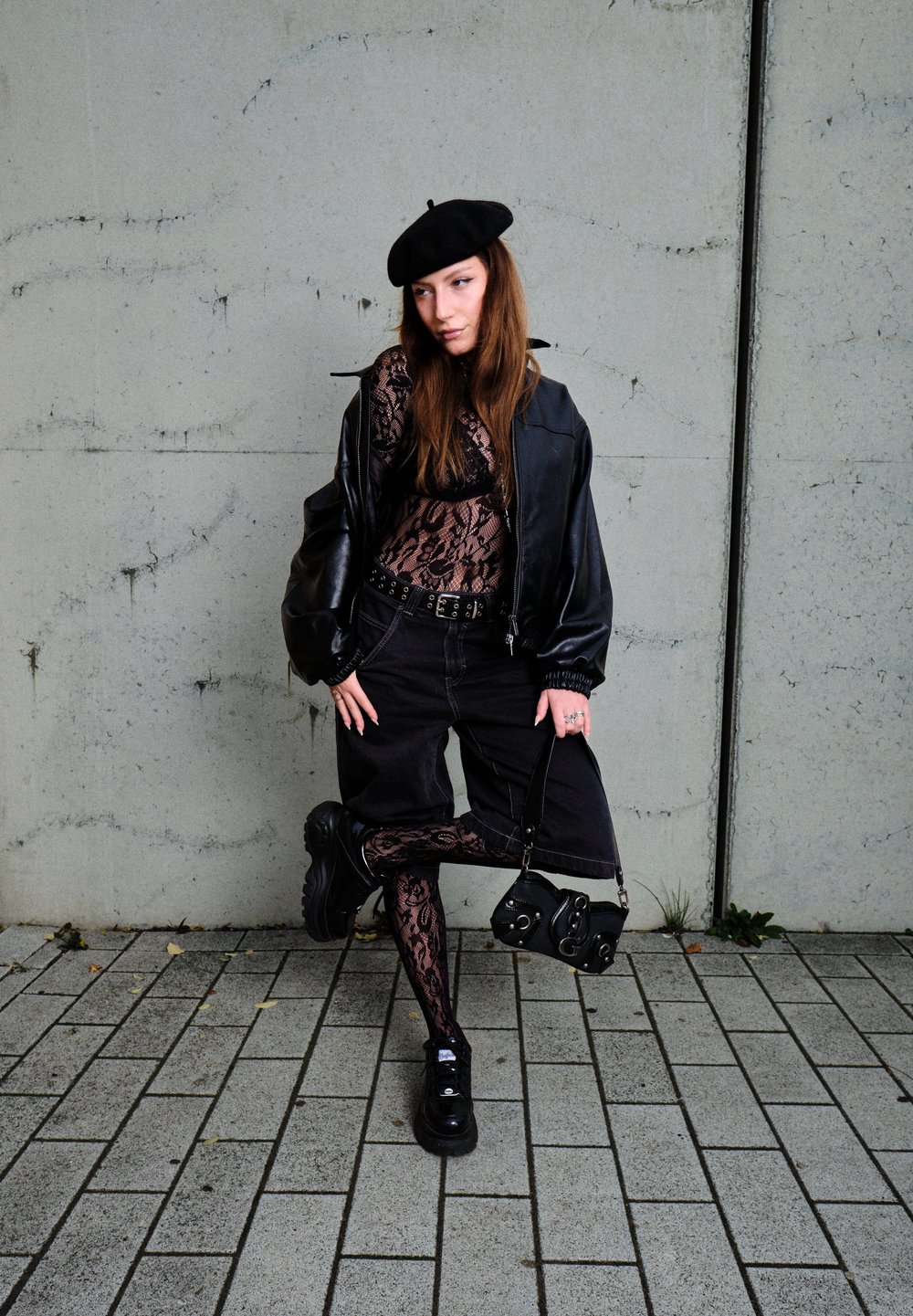 Μαύρη δερμάτινη τζάκετ bomber πάνω από ένα κορμάκι από δαντέλα, συνδυασμένο με μαύρες βερμούδες και κολάν από δαντέλα. Μια μαύρη μπερέ και παπούτσια με χοντρή σόλα ολοκληρώνουν την εμφάνιση.