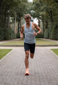 Jooksja helesinises tanktopis ja mustades lühikestes pükstes, jalas oranžid jooksujalatsid ja randmelt nutikell, tellistest teel, mille ääres on rohelus.