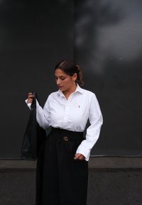 Camicia bianca a bottoni con logo, abbinata a una gonna nera a vita alta con fibbia dorata. Borsa nera in mano.