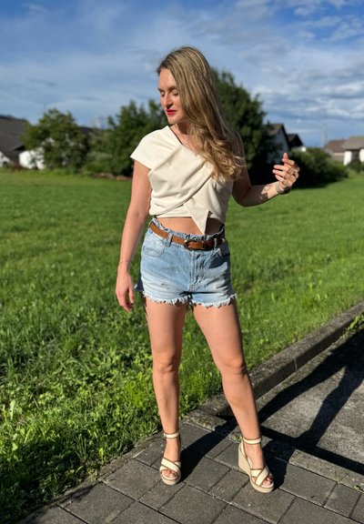 Top beige cruzado con mangas cortas, shorts de denim de cintura alta con dobladillo deshilachado y sandalias de cuña beige sobre un camino pavimentado en un área de césped.