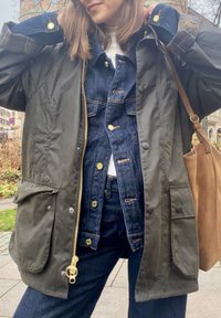 Ciemnozielona, wodoodporna kurtka z złotym zamkiem i kieszeniami, nałożona na niebieską kurtkę dżinsową i białą koszulę. Widoczna brązowa torba na ramię.