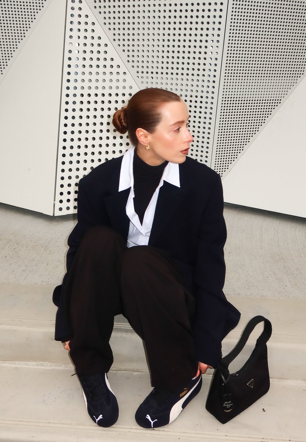 Svart oversized blazer, vit skjorta, svart turtleneck, lösa svarta byxor och marinblå sneakers. Liten svart handväska med logotyp. Geometrisk bakgrund.