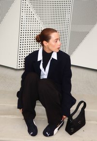 Svart oversized blazer, vit skjorta, svart turtleneck, lösa svarta byxor och marinblå sneakers. Liten svart handväska med logotyp. Geometrisk bakgrund.
