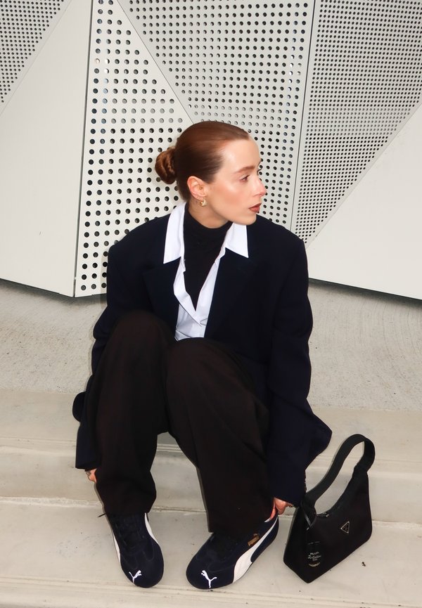 Blazer nero oversize, camicia bianca, dolcevita nero, pantaloni neri larghi e sneakers blu navy. Piccola borsa nera con logo. Sfondo geometrico.