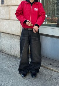 Κόκκινο ζιβάγκο μπουφάν με άσπρη γραφή, μαύρη κουκούλα από κάτω, συνδυασμένο με φαρδιά μαύρα τζιν και γυαλιστερές μαύρες μπότες.