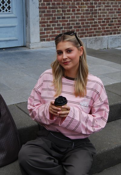 Sudadera holgada de rayas en rosa claro con mangas largas y detalle de texto, combinada con pantalones de pierna ancha en gris oscuro. Sosteniendo una taza de café.
