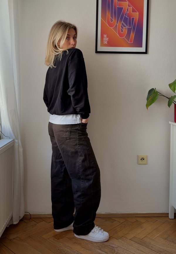 Junge Frau mit blondem Haar, die schwarze Hose, schwarzen Pullover und weiße Sneakers trägt, steht mit den Händen in den Taschen neben einer Wand mit einem farbenfrohen gerahmten Plakat.