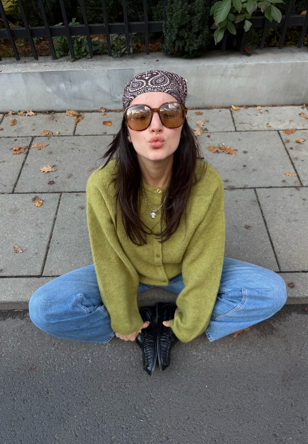 Une femme portant des lunettes de soleil, un foulard à motifs sur la tête, un pull vert et un jean est assise en tailleur sur le trottoir et pince les lèvres face à l’appareil photo.