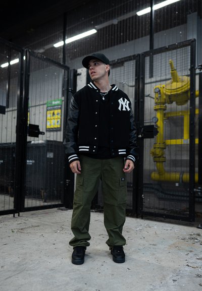 Chaqueta tipo letterman en blanco y negro con mangas de cuero, combinada con camiseta negra y pantalones cargo verdes. Zapatillas negras completan el atuendo.