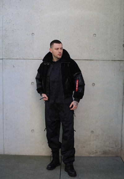 Cazadora bomber de cuero negro y borreguillo con cuello oversized, combinada con overoles de utilidad negros y botas negras con cordones.
