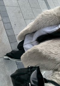 Béžový umělý kožich s širokými rukávy přes šedou mikinu na zip a černé kalhoty s bílými pruhy. Černá kabelka s kovovými detaily.