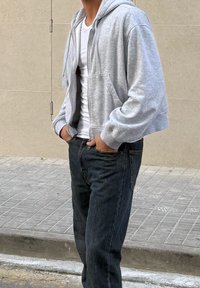 Oseba v svetlo sivi jopi s kapuco na zadrgo, oblečena čez belo majico, z rokami v žepih, stoji na tlakovanem hodniku ob bež steni.