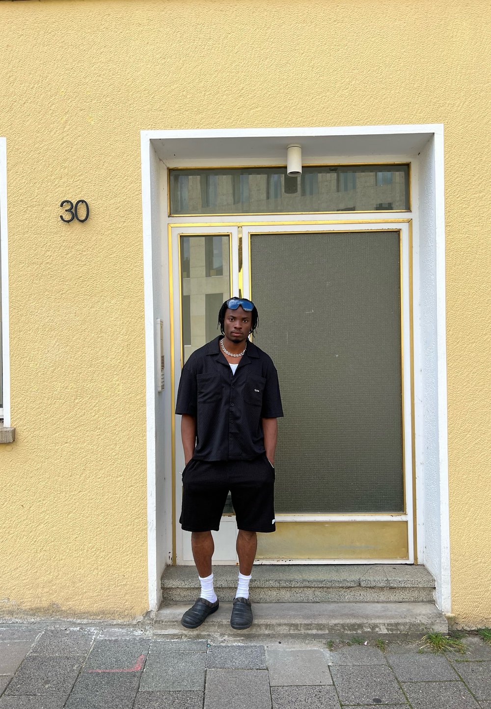 Mustamine lühikeste käistega nööpidega särgid, mustad lühikesed püksid, valged sokid ja mustad libisemisjalanõud. Seisab kollase seina ukse juures, millel on klaaspaneel.