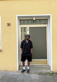 Mustamine lühikeste käistega nööpidega särgid, mustad lühikesed püksid, valged sokid ja mustad libisemisjalanõud. Seisab kollase seina ukse juures, millel on klaaspaneel.