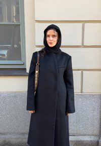 Juodas vilnonis paltas su aukštu apykakliu, turintis užslėptą priekinę dalį, ilgomis rankovėmis ir aptemptą siluetą. Tekstūruota megzta gobtuvas ir ruda rankinė su raštuotu dirželiu.