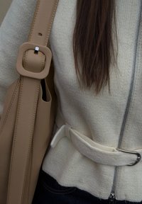 Gros plan sur un pull en maille crème à fermeture éclair avec ceinture, cheveux foncés, et sac à bandoulière en cuir beige avec boucle carrée.