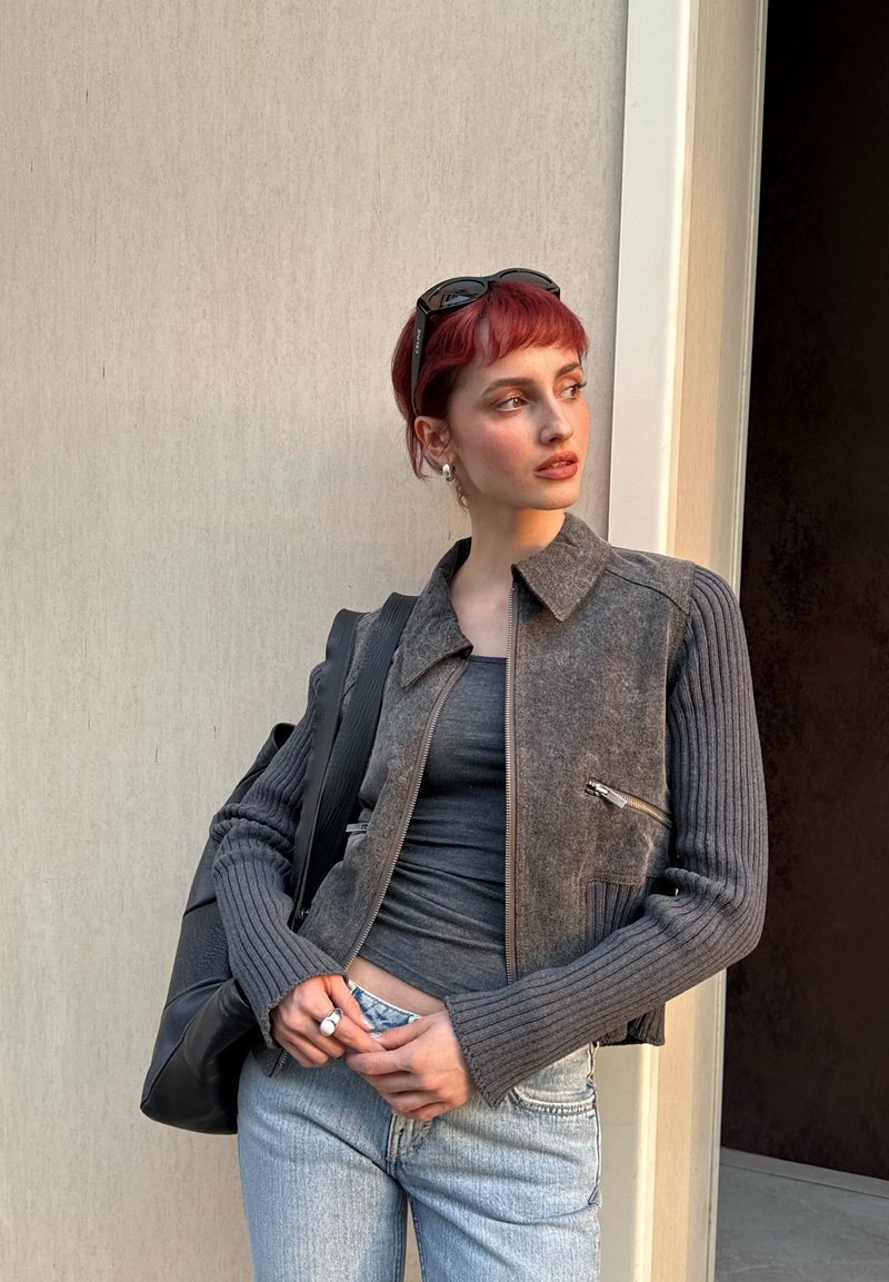 Jeune personne aux cheveux rouges courts portant une veste grise, un débardeur foncé, un jean clair et des lunettes de soleil sur la tête, debout contre un mur clair.