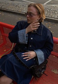 Τζάκετ από τζιν με αντίθεση στους φωτεινούς μπλε μανίκους, φορεμένο πάνω από ένα φόρεμα τζιν. Συνδυάζεται με μια μαύρη ζώνη χιαστί και πολλούς δακτυλίους. Οι καφέ γυαλιά ηλίου συμπληρώνουν την εμφάνιση.