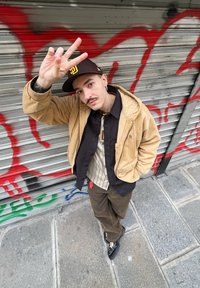 Ταν μπουφάν με κουκούλα πάνω από μαύρο πουκάμισο, ριγέ μπεζ T-shirt, καφέ παντελόνι και μαύρα παπούτσια. Χέρι υψωμένο με το σύμβολο της ειρήνης μπροστά από γραφικά.