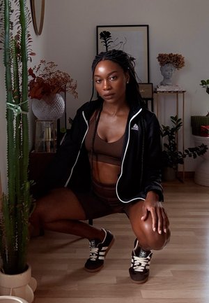 Chaqueta negra con cremallera y acentos blancos, sujetador deportivo marrón y pantalones cortos, zapatillas negras con rayas blancas, y un fondo texturizado con vegetación.