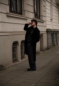 Manteau long noir, pull vert foncé avec logo, pantalon large gris, chaussures noires. Une personne se tient devant un bâtiment, ajustant ses lunettes de soleil.