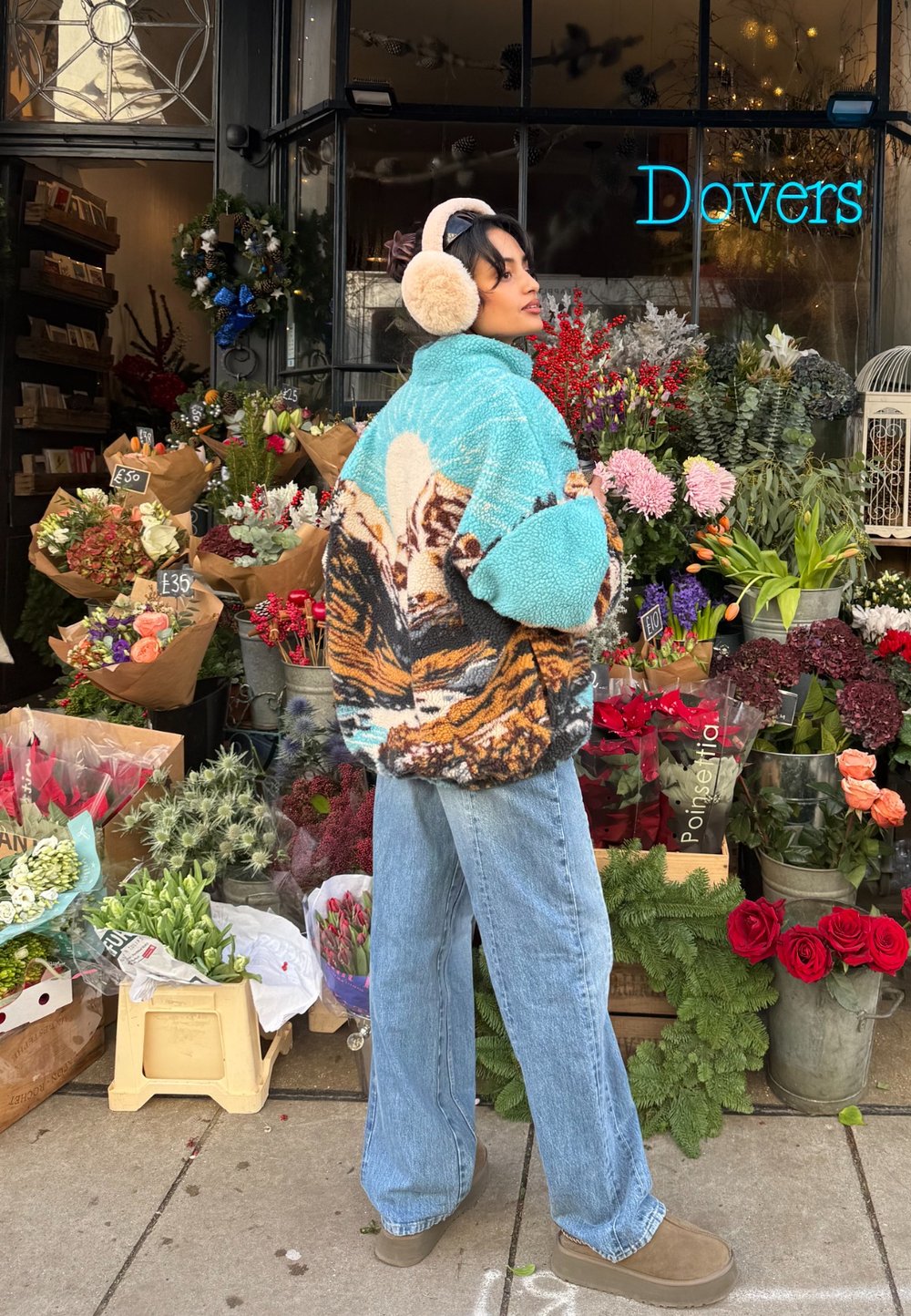 Fuzzy turquoise jas met een berglandschap, gecombineerd met lichtblauwe jeans en bruine instappers. Omringd door kleurrijke bloemstukken.
