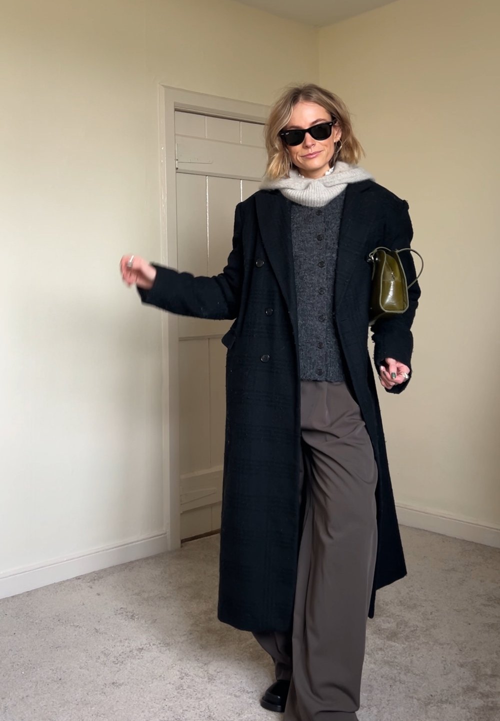 Cappotto lungo nero sopra un maglione grigio con bottoni, abbinato a pantaloni beige a gamba larga. Gli accessori includono occhiali da sole neri e una piccola borsa verde.