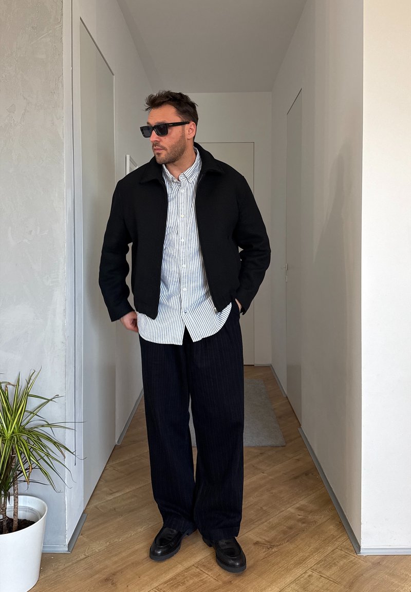 Homme portant des lunettes de soleil noires, une veste noire, une chemise à rayures boutonnée et un pantalon large noir, debout dans un couloir étroit à côté d'une plante en pot.
