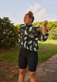 Shorts en vélo noirs associés à une veste ample et à motifs, présentant des formes géométriques vertes sur un fond marine. Cadre extérieur avec de la verdure.