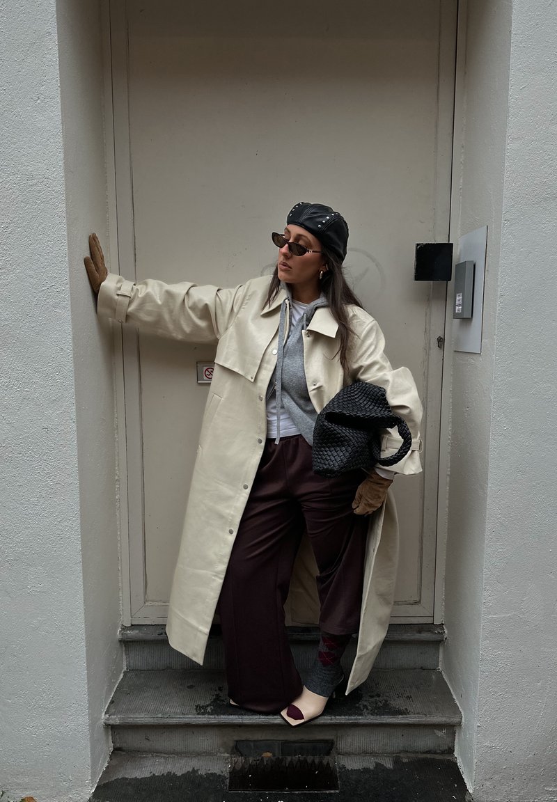Manteau oversize couleur crème, sweat à capuche gris, pantalon large bordeaux, chaussettes à motifs, gants, sac à main noir, lunettes de soleil foncées et casquette noire.