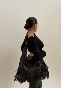 Kvinna med svart hår i en låg knut bär en svart off-shoulder topp och håller en stor svart fransad läderväska över axeln mot en enfärgad vägg.